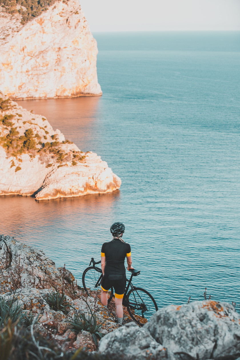 Una mujer está de pie junto a un acantilado con su bicicleta y mira hacia el mar.